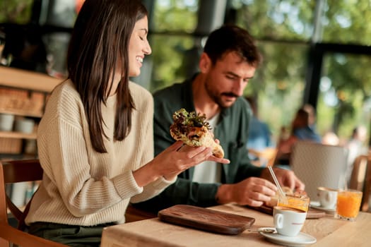 Couple shares laughter and delicious food while sitting at a wooden table in a bright cafe, surrounded by greenery and other patrons enjoying their meals