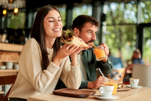Couple shares laughter and delicious food while sitting at a wooden table in a bright cafe, surrounded by greenery and other patrons enjoying their meals