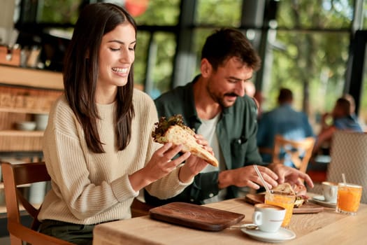 Couple shares laughter and delicious food while sitting at a wooden table in a bright cafe, surrounded by greenery and other patrons enjoying their meals
