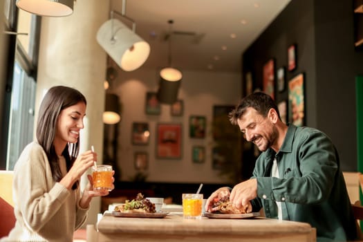 Couple shares laughter and delicious food while sitting at a wooden table in a bright cafe, surrounded by greenery and other patrons enjoying their meals