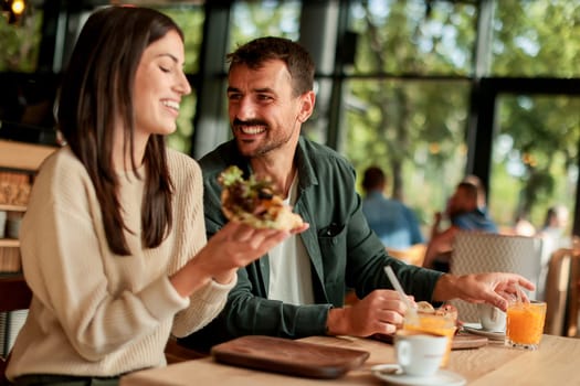Couple shares laughter and delicious food while sitting at a wooden table in a bright cafe, surrounded by greenery and other patrons enjoying their meals