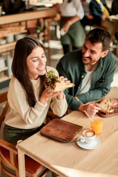 Couple shares laughter and delicious food while sitting at a wooden table in a bright cafe, surrounded by greenery and other patrons enjoying their meals