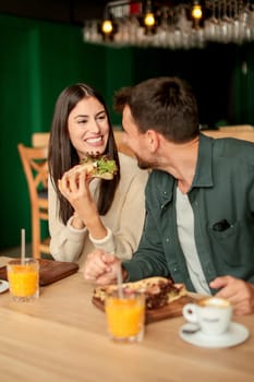 Couple shares laughter and delicious food while sitting at a wooden table in a bright cafe, surrounded by greenery and other patrons enjoying their meals