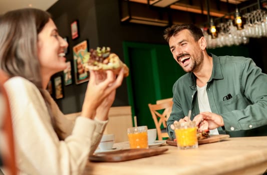 Couple shares laughter and delicious food while sitting at a wooden table in a bright cafe, surrounded by greenery and other patrons enjoying their meals