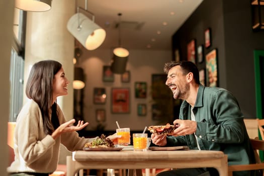 Couple shares laughter and delicious food while sitting at a wooden table in a bright cafe, surrounded by greenery and other patrons enjoying their meals