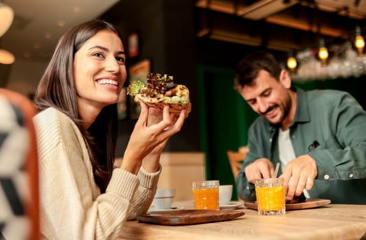 Couple shares laughter and delicious food while sitting at a wooden table in a bright cafe, surrounded by greenery and other patrons enjoying their meals
