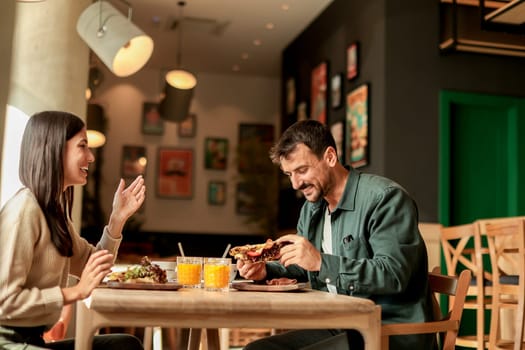 Couple shares laughter and delicious food while sitting at a wooden table in a bright cafe, surrounded by greenery and other patrons enjoying their meals