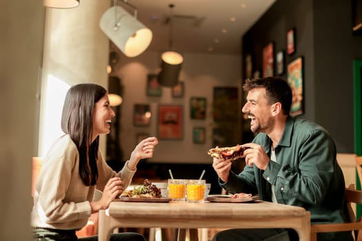Couple shares laughter and delicious food while sitting at a wooden table in a bright cafe, surrounded by greenery and other patrons enjoying their meals