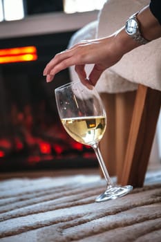 girl with watch on hand holds glass of wine with two fingers, fireplace burning in background in modern interior, romantic atmosphere of relaxation. Warm atmosphere