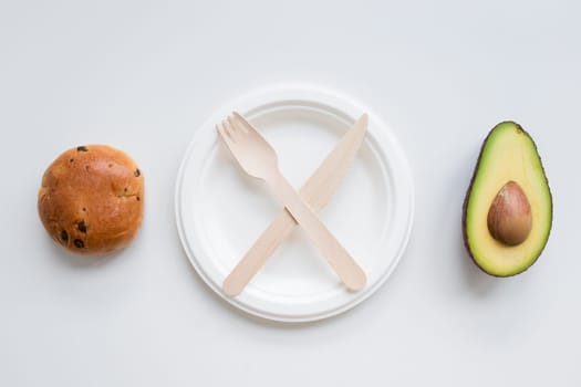 Minimalist flat lay with knife, fork, avocado, and bread roll on white background.
