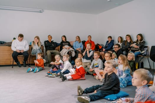 Little children with parents sit on cushions on the floor and chairs against the wall and watch a performance in a kindergarten. High quality photo