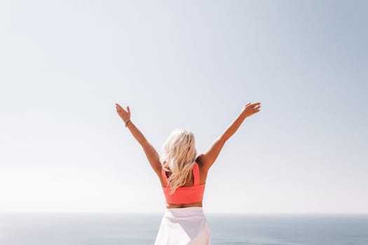 A woman in a pink top and white skirt is standing on a cliff overlooking the ocean. She is smiling and she is happy