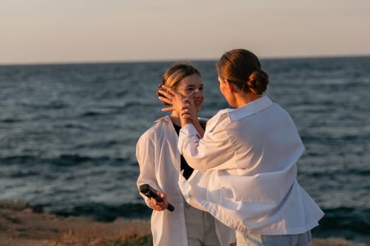 Two women, wearing white shirts, stand by the ocean at sunset. One woman holds a microphone and touches the other's face.