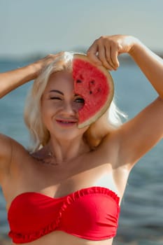 Blonde woman in red bikini playfully holds a watermelon slice to her face, partially obscuring her features. Sunny beach backdrop.