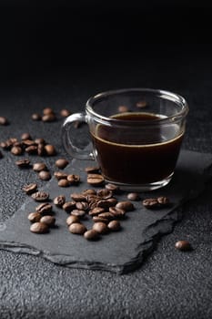 Dark coffee in a glass coffee cup, black espresso and roasted coffee beans pilled on black table, close up. Vertical banner with copy space
