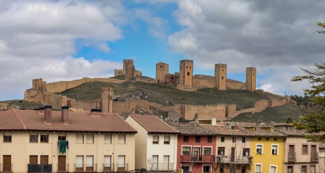 A majestic medieval castle on a hill overlooks colorful houses, beneath a partly cloudy sky.