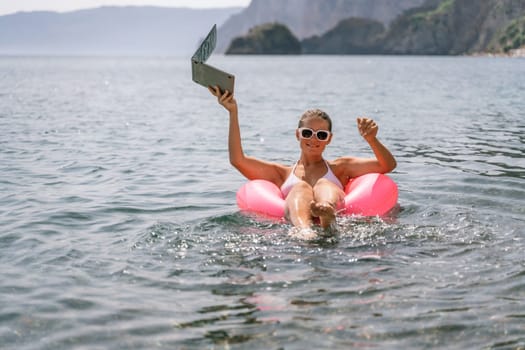 A woman is floating in a pink inflatable raft with a laptop in her hand. She is smiling and she is enjoying her time in the water