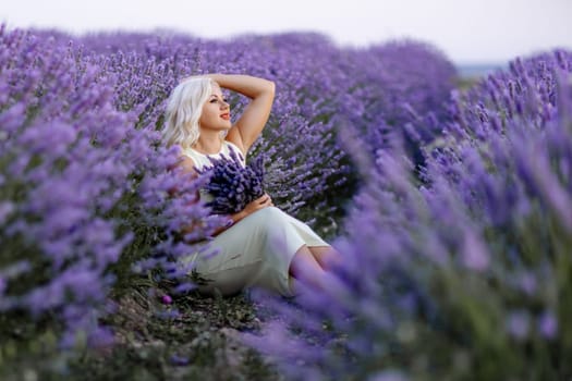 Blonde woman poses in lavender field at sunset. Happy woman in white dress holds lavender bouquet. Aromatherapy concept, lavender oil, photo session in lavender.