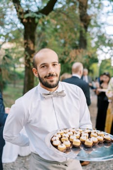 Smiling waiter with cupcakes on a tray standing in the park. High quality photo