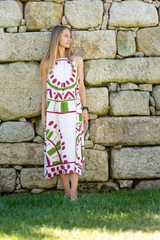Young woman is posing in a summer dress while leaning against a rustic stone wall