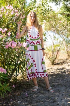 Young blonde woman is posing in a botanical garden, wearing a long colorful dress and touching oleander flowers