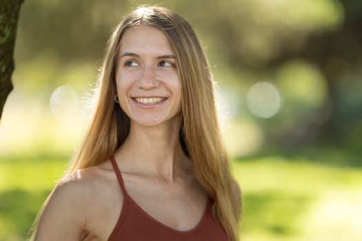 Young woman with long blond hair is smiling and looking away while standing in a park