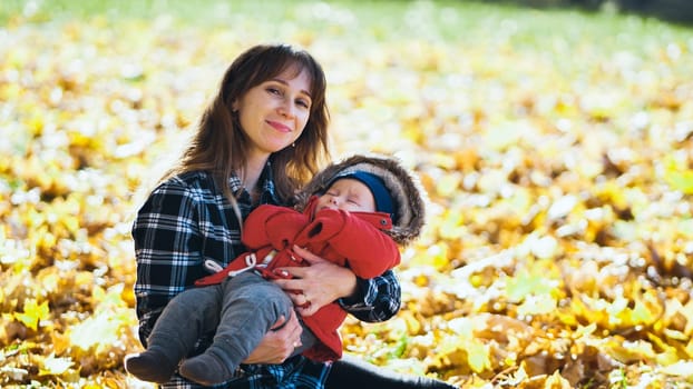 Mother cuddling peacefully sleeping infant amid golden autumn foliage, warm sunlight filtering through colorful falling leaves