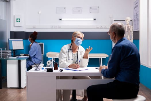 Retired man sits at desk, discussing healthcare details with old female physician in lab coat, reviewing treatment notes. Elderly doctor uses clipboard to share medical results with pensioner patient.