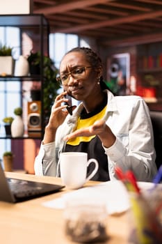 Remote worker in home office upset by bad news received during phone call. Annoyed african american woman receiving frustrating telephone call on smartphone from job coworker