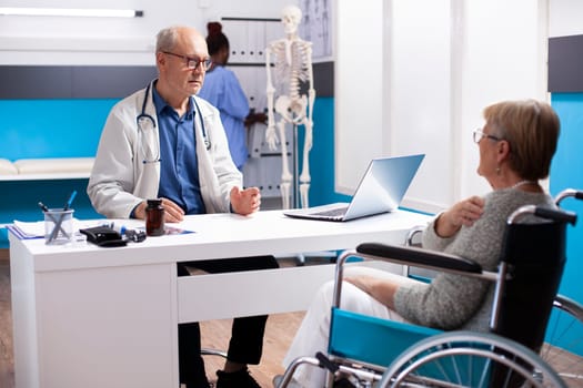 Senior male doctor with laptop on table is speaking to female pensioner patient about her recovery treatment process. Retired physician engages with old woman in wheelchair about her healthcare plan.