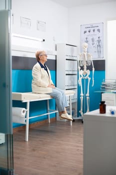 Elderly woman in casual clothing seated in hospital room ready for health consultation. Caucasian senior lady sitting on bed and waiting in doctors office for a medical checkup.