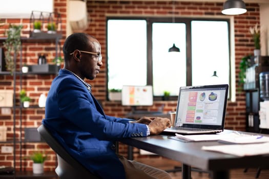 African american man focused on reviewing financial charts displayed on digital device in startup office. Male analyst with glasses, sitting at desk and working on his laptop, doing company research.