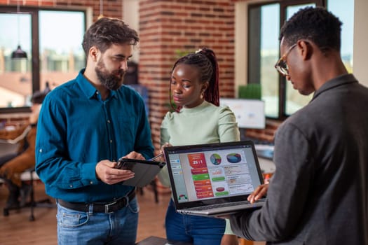 Businessman and woman reviewing online documents on tablet as black man checking business analytics on his laptop. Male and female coworkers using digital devices in brick wall office.