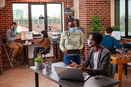 Female employee holding tablet and receiving instructions from male manager seated at desk in brick wall office. African american entrepreneurs with face mask, having a discussion in modern workspace.