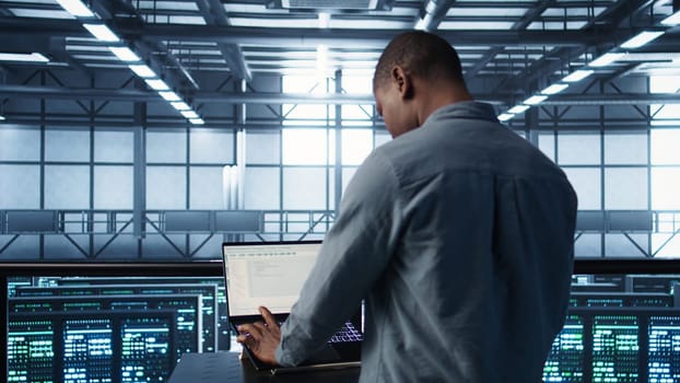 Computer scientist in server room coding on laptop, developing disaster recovery plans. African american man in data center testing failover scenarios using notebook, camera B