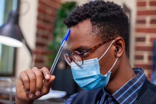 Closeup of black man wearing face mask and touching his forehead with a pen in startup office. African american male employee tapping his head and listening to music with earbuds in brick wall room.