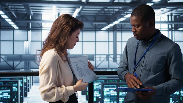 Team of IT professionals looking over data center checkup checklist paperwork. African american man and colleague collaborating in server farm reading documentation files, camera B