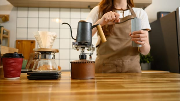Cropped shot of woman in an apron grinding coffee beans by hand beside a pour over dripper.