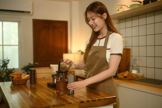 Young woman pours coffee beans into a grinder on a wooden countertop.