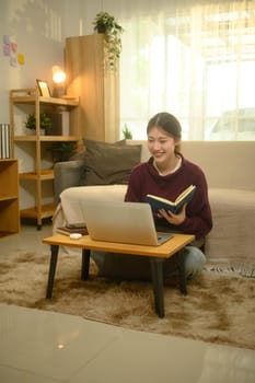 Young female student holding notebook and using laptop in living room.