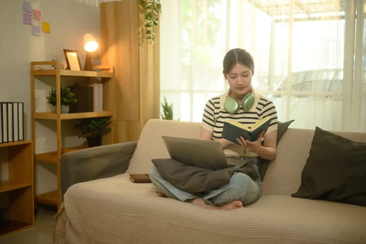 Female student reading a book with laptop on her lap sitting on couch at home.