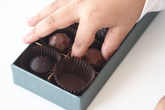 A hand selects chocolates from an open box on a white surface.