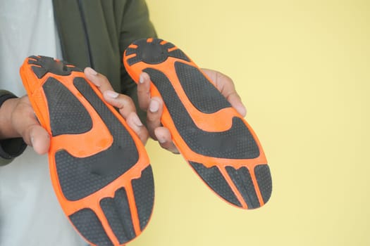 Two orange and black shoe soles are displayed by a person in front of yellow.