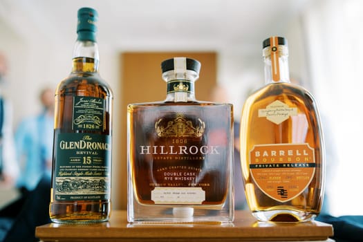 Bottles of whiskey stand on the table in front of the men. Caption: Glendronach Revival, Hillrock, Barrell Bourbon. High quality photo