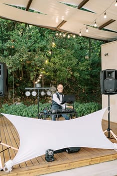 DJ sits on a chair at the console on a stage under cover in the garden. High quality photo