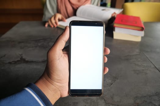 A person holds a blank smartphone while studying with books nearby.