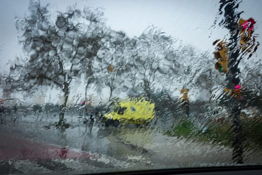 A view through a rain-soaked window showcases blurred urban scenery, capturing the mood of a rainy day with vibrant colors blending amidst the downpour.