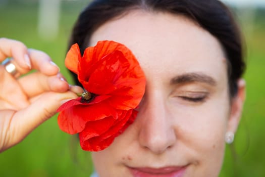 A young woman gently holds a vibrant red poppy flower to her closed eye, creating a serene and beautiful moment outdoors.