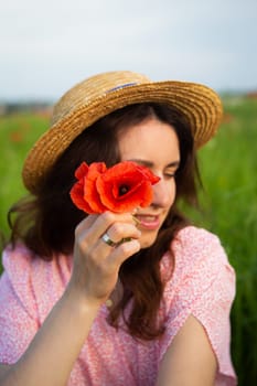 A young woman in a straw hat holds a vibrant red poppy flower near her face in a sunny field.
