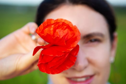 A smiling woman playfully covers her eye with a vibrant red poppy flower, enjoying a sunny day outdoors.